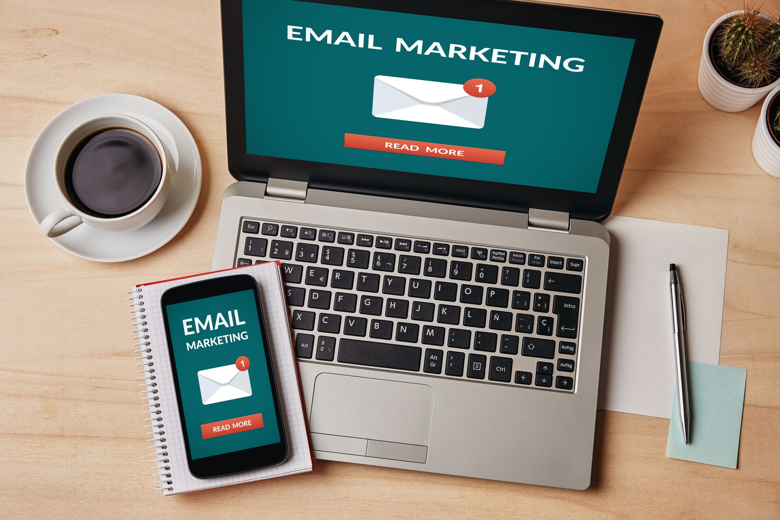 A laptop and smartphone showing an "Email Marketing" screen sit on a wooden desk, surrounded by a coffee cup, pen, and notepad. Perfectly poised to leverage these tools, small business owners can seamlessly boost their communication strategies.