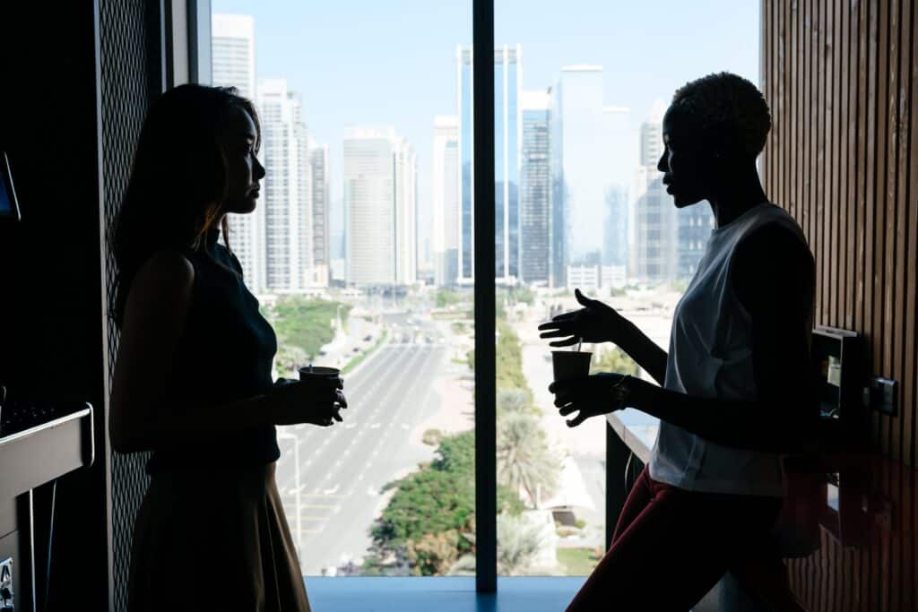 Two people having a conversation by a window overlooking a cityscape.