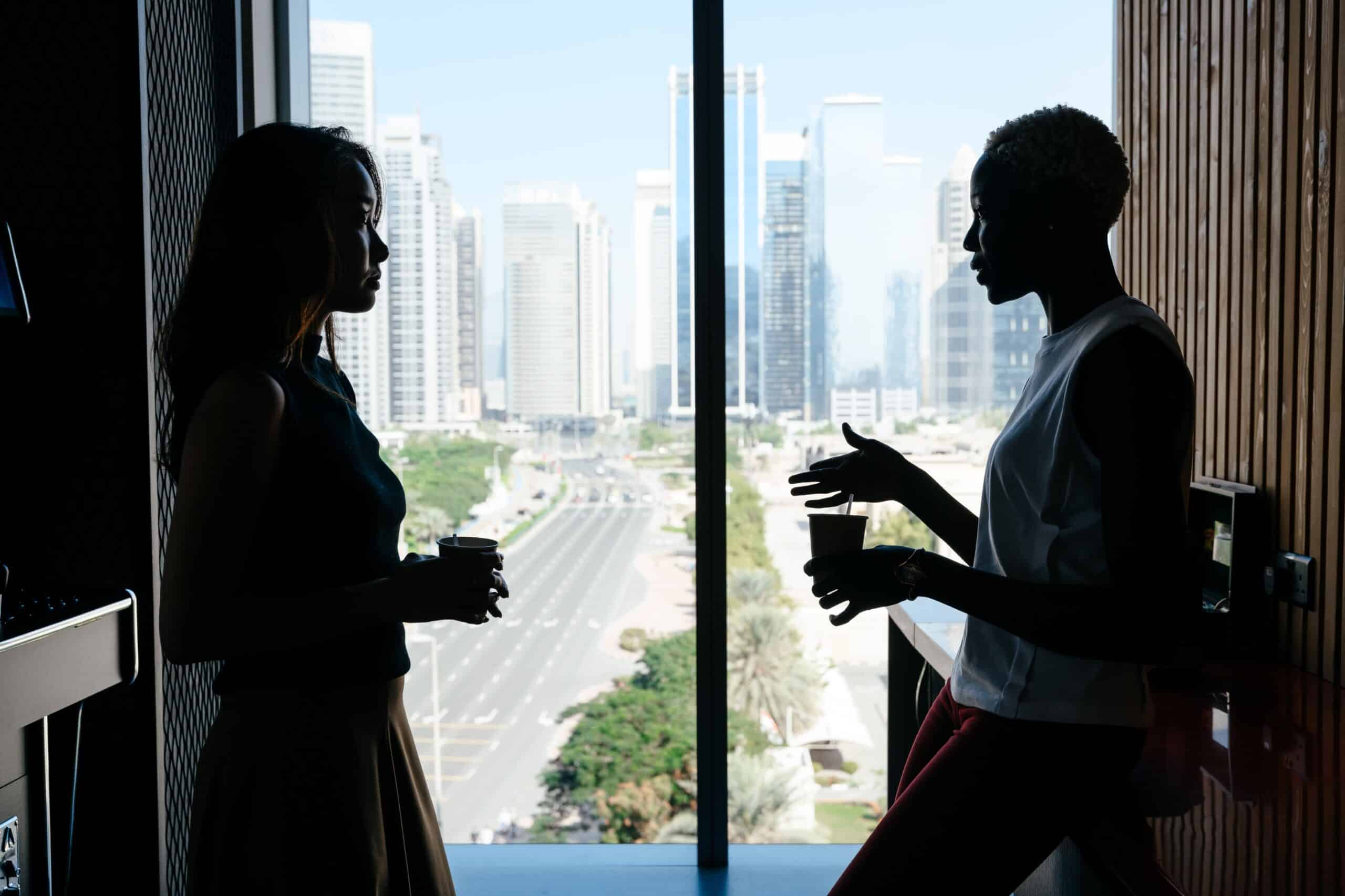 Two people are silhouetted against a large window overlooking a cityscape, holding cups and engaged in conversation.