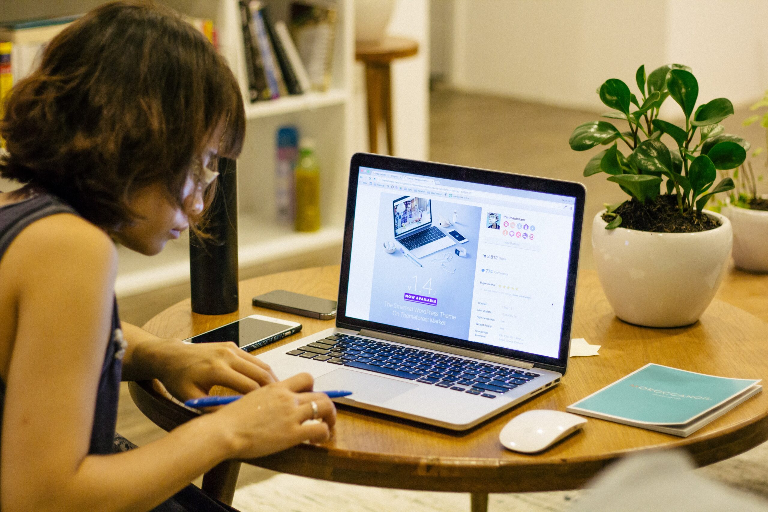 Person working on a laptop at a wooden table, surrounded by a notebook, smartphone, and a potted plant.