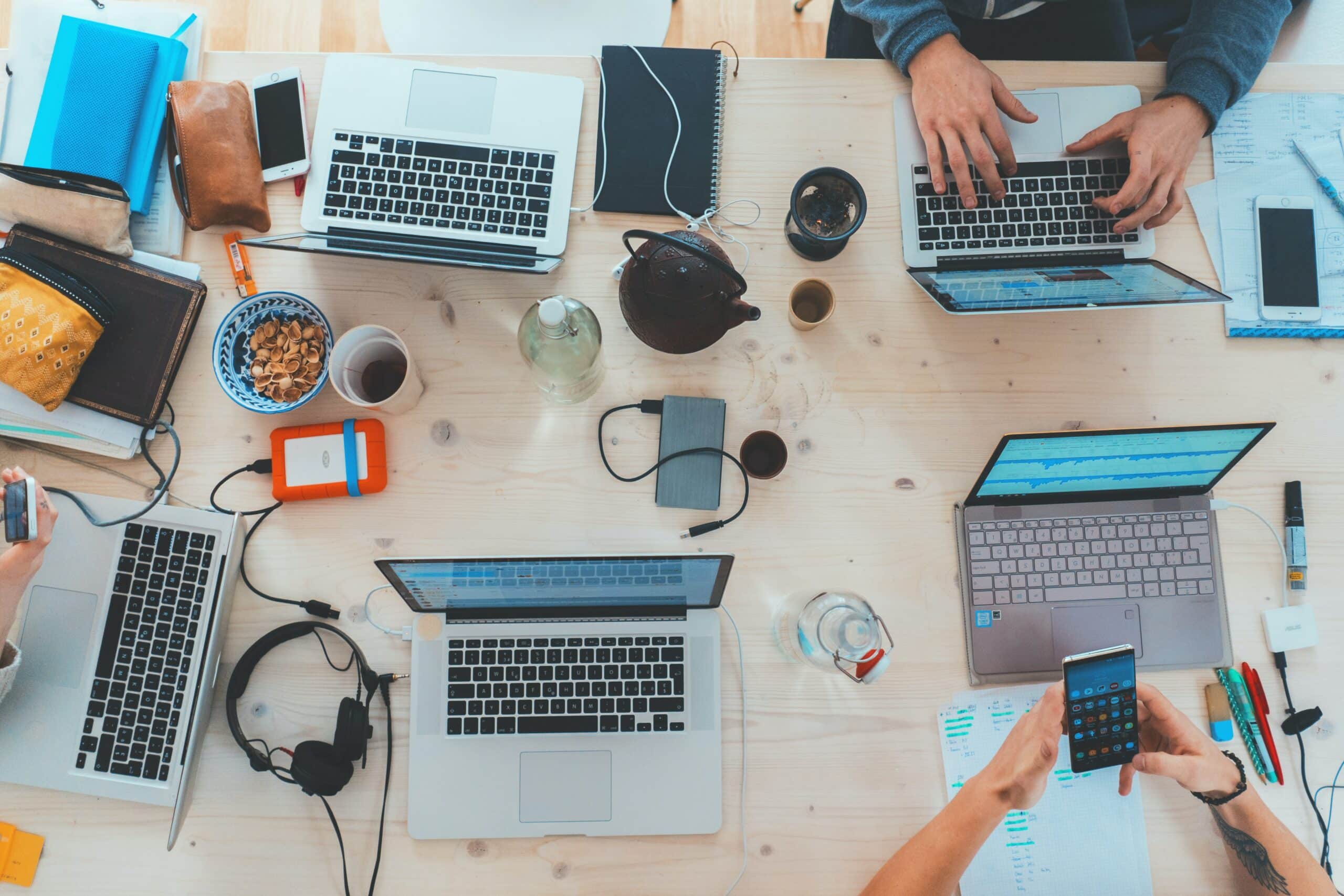 Aerial view of a desk with laptops, notebooks, phones, and various items. People are working and using electronic devices, with a bowl of snacks and drinks visible.