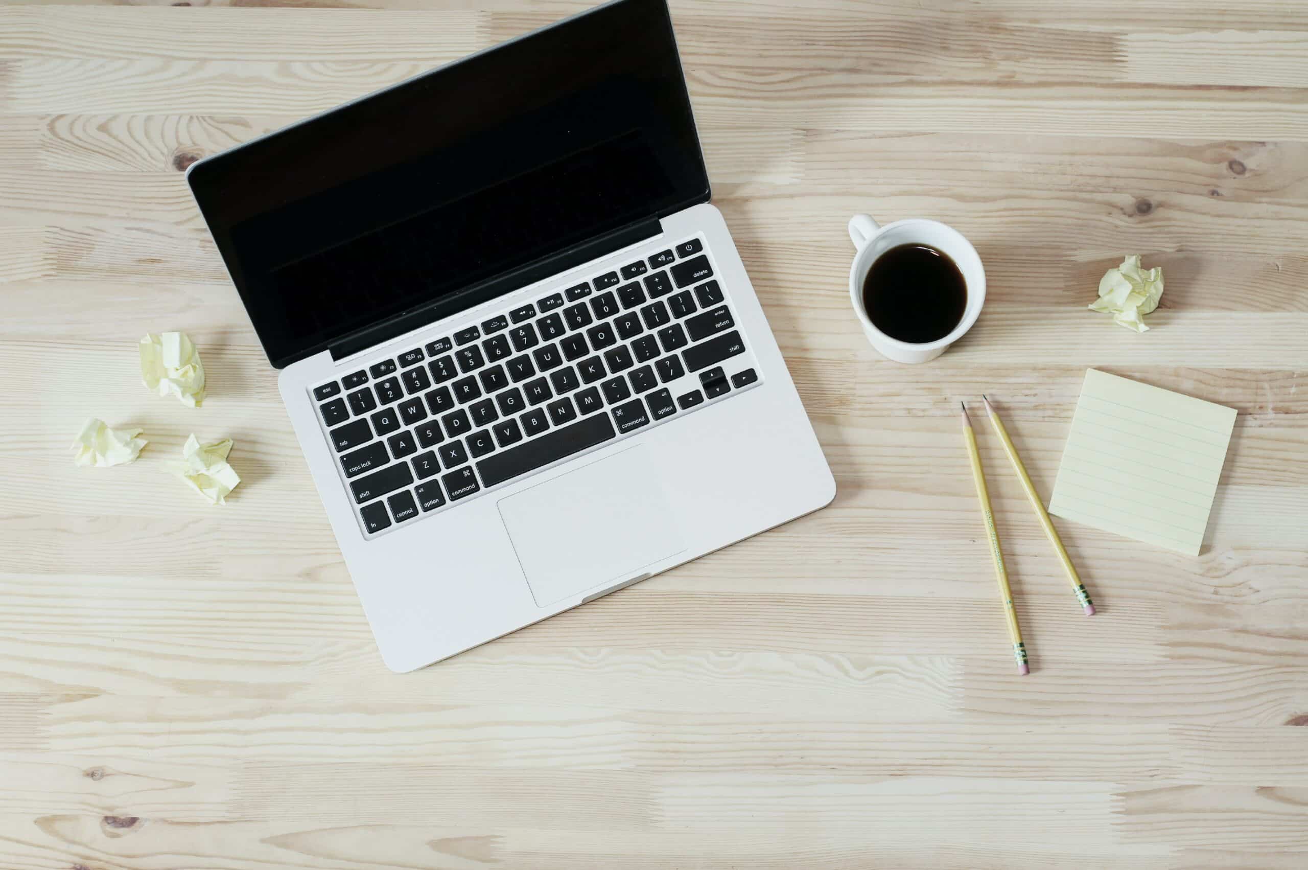 A laptop on a wooden desk with a cup of coffee, two pencils, crumpled paper, and a notepad.