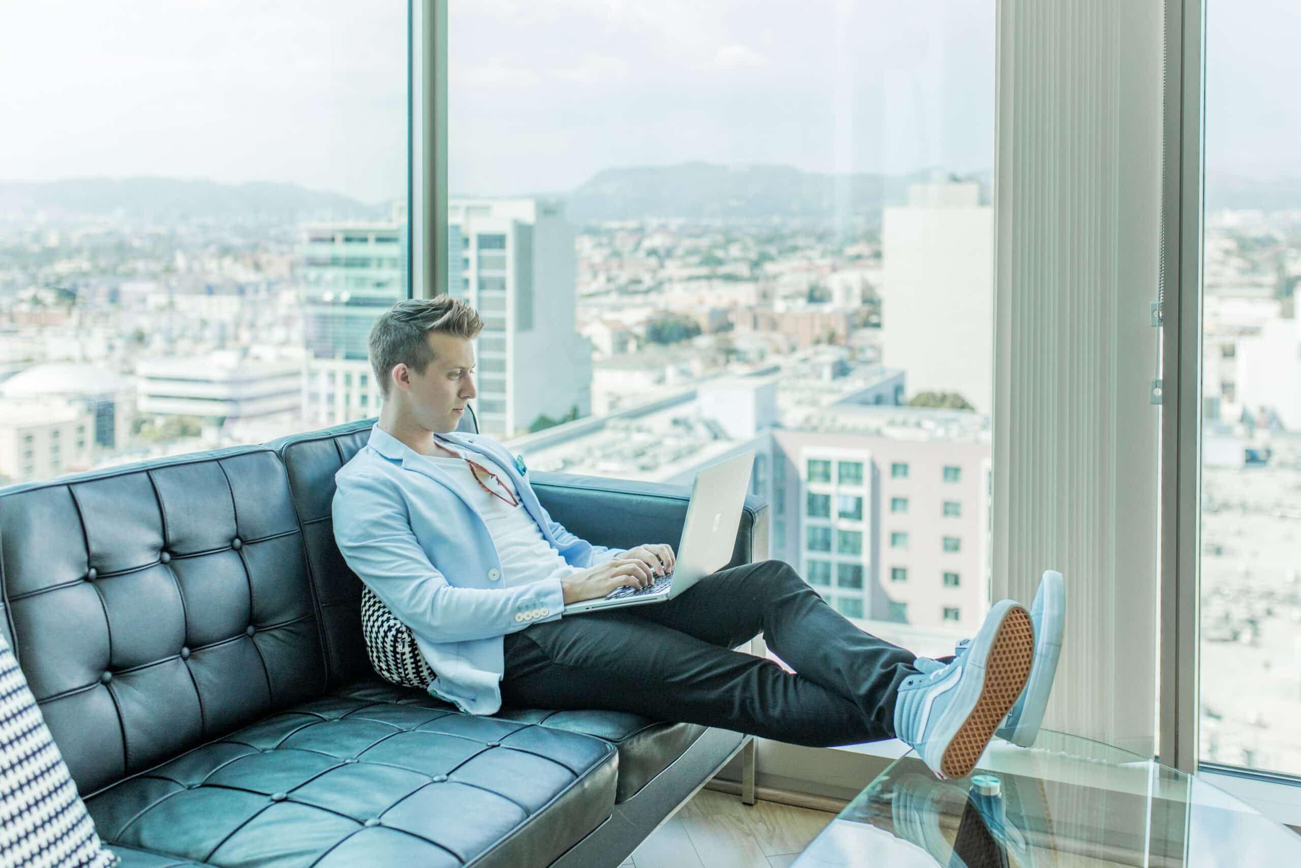 A person sits on a black leather couch, using a laptop, with a cityscape visible through large windows in the background.