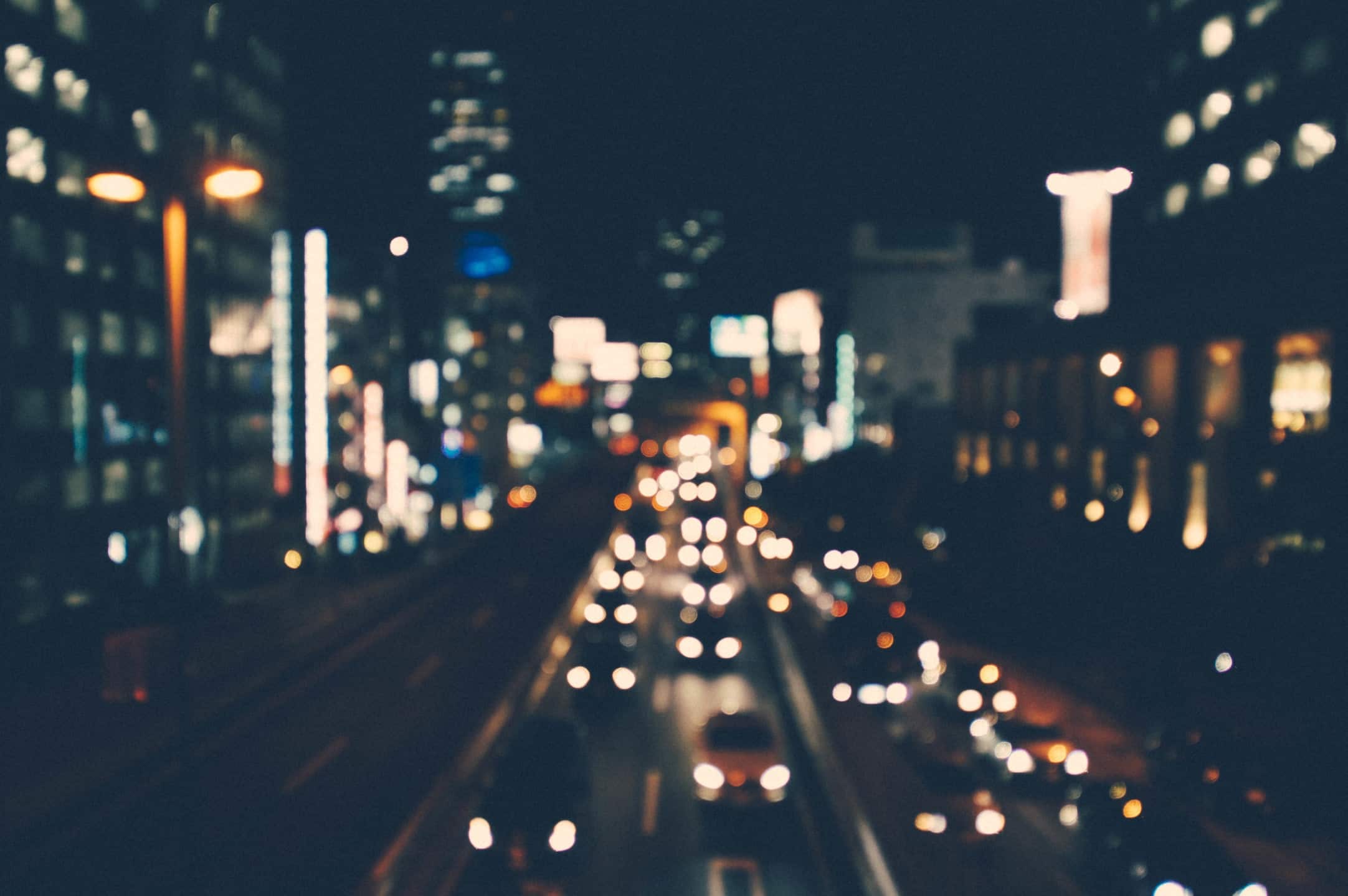 Blurred image of city street at night with illuminated buildings and moving car lights.