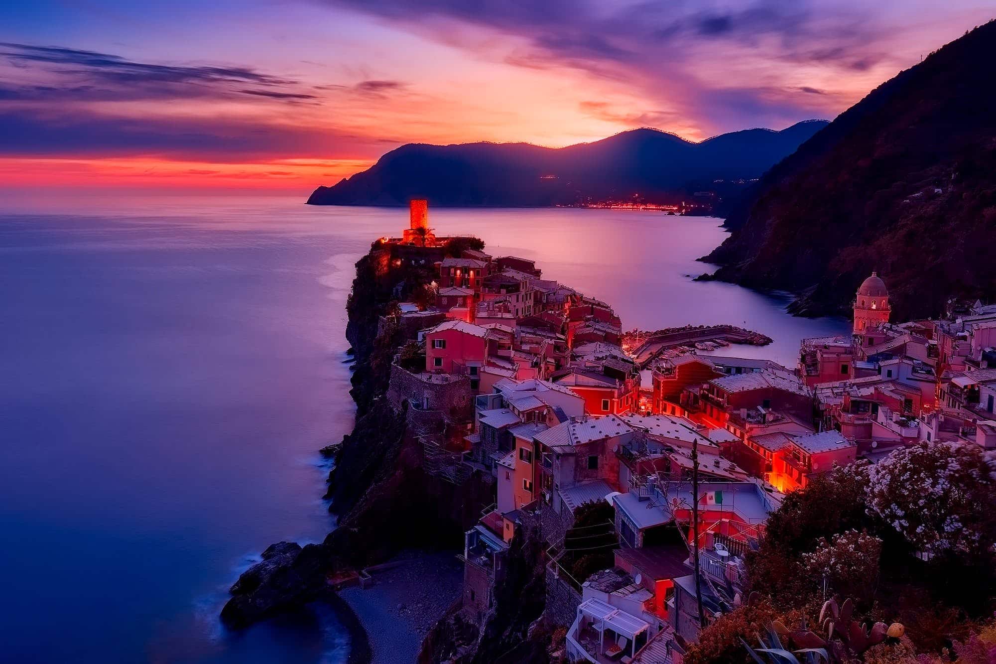 Coastal village on a cliff at sunset, with colorful houses and illuminated streets. The sea is calm, and the sky is a gradient of orange and purple hues.