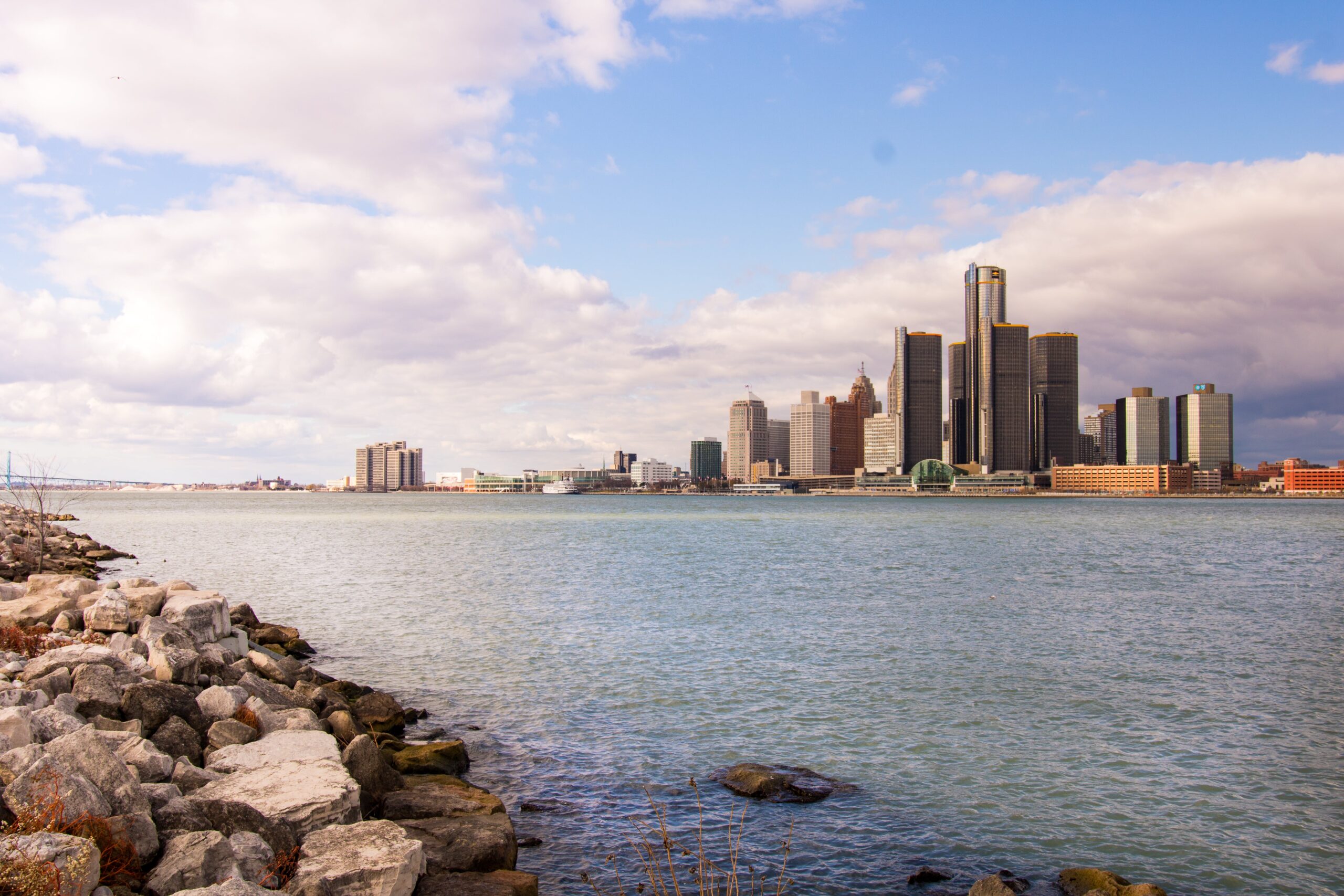 Skyline of a city with tall buildings across a body of water, rocky shoreline in the foreground, under a partly cloudy sky.