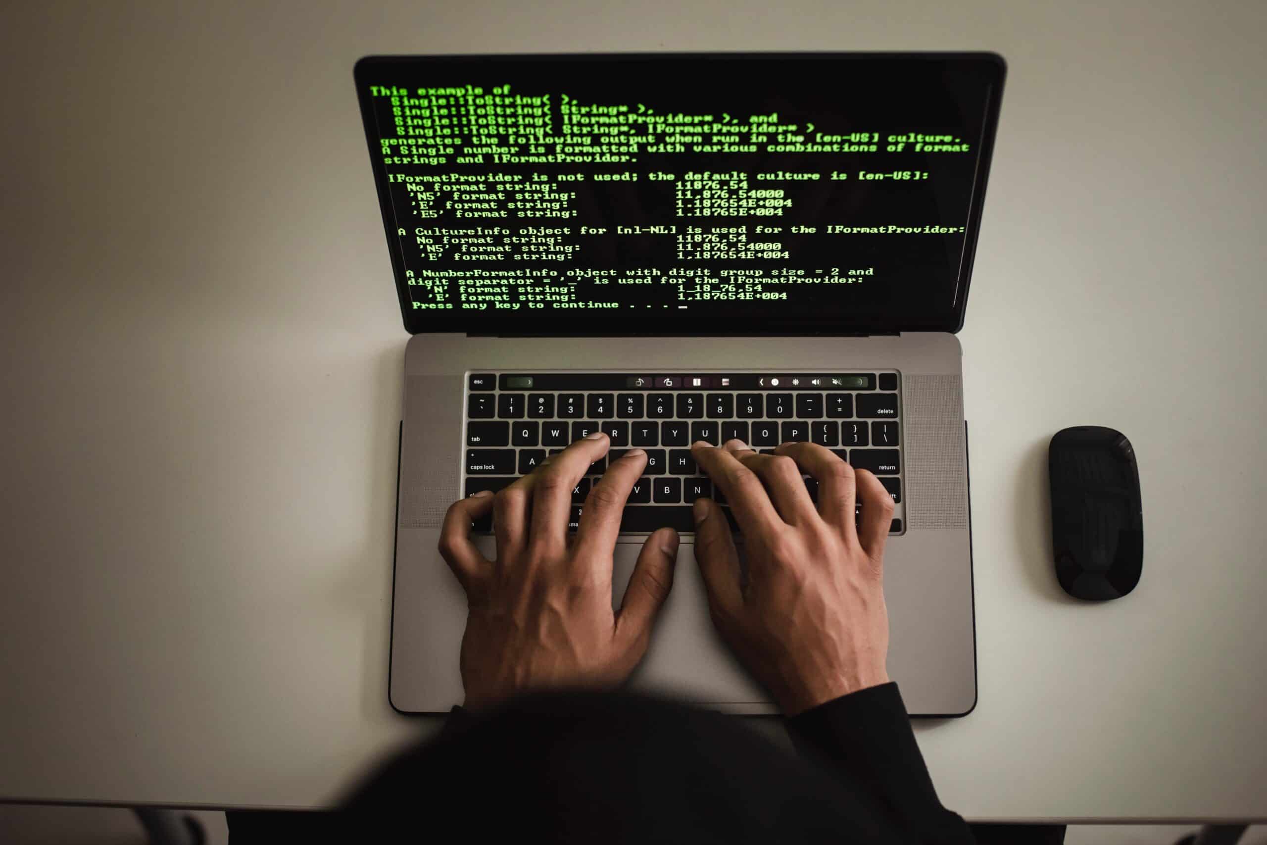 Person typing on a laptop displaying code in a dark-themed text editor, with a black mouse beside it on a white surface.