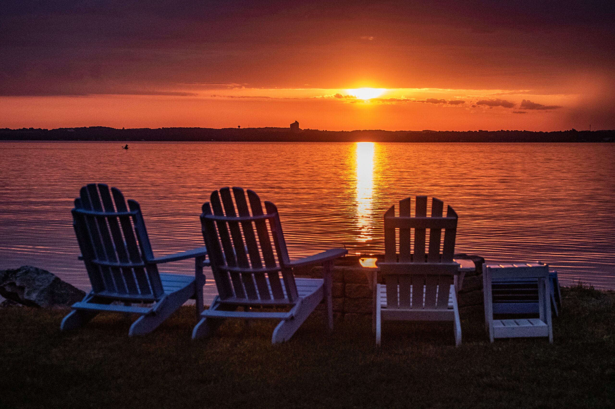 Four Adirondack chairs face a lake under a glowing sunset, with the sun's reflection shimmering on the water.