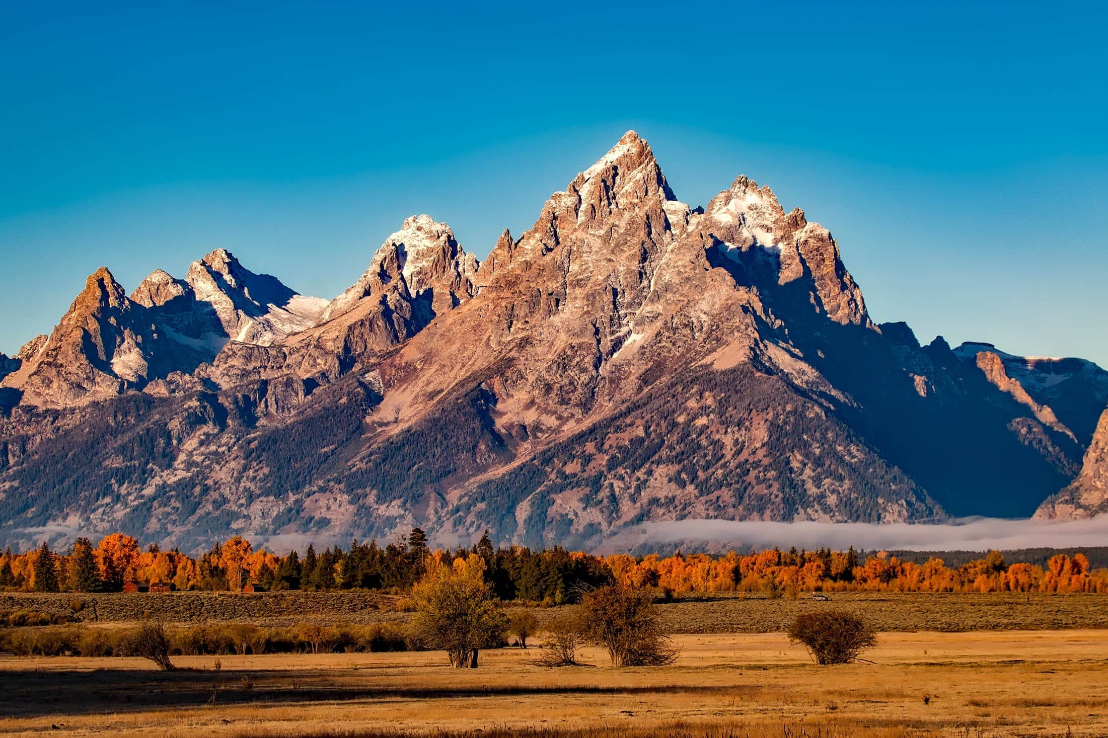 Mountain range with rocky peaks under a clear blue sky, surrounded by an autumnal landscape with trees and open fields in the foreground.