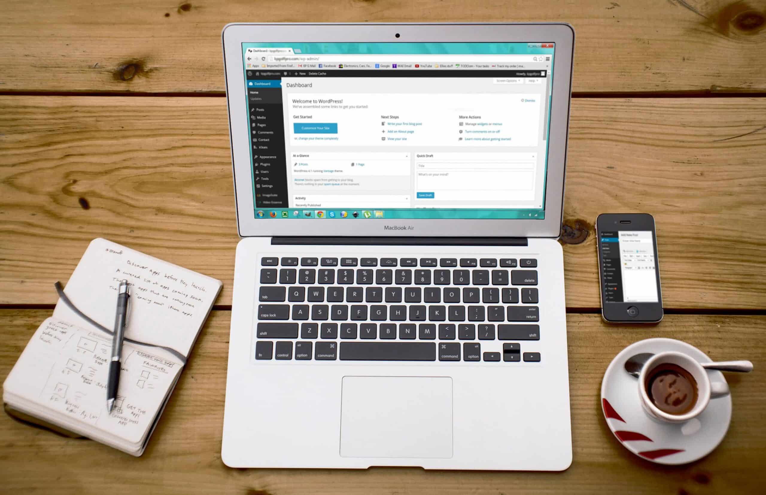 Open laptop displaying a dashboard, next to a notebook, smartphone, and a cup of coffee on a wooden table.