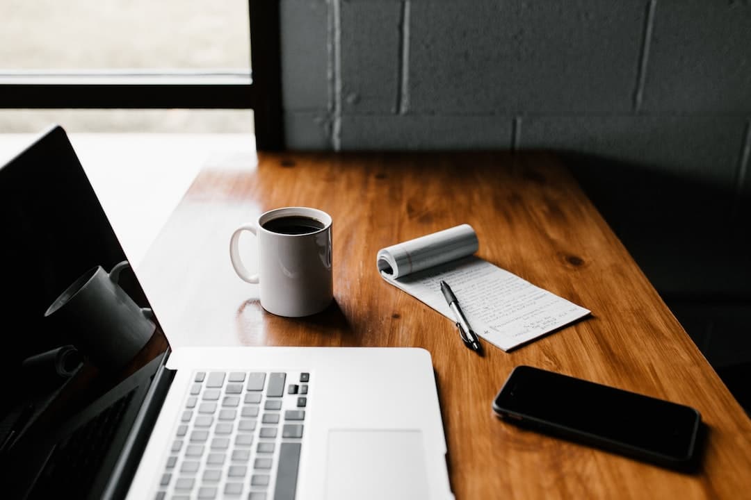 A wooden desk with a laptop, a cup of coffee, a notepad with a pen, and a smartphone.