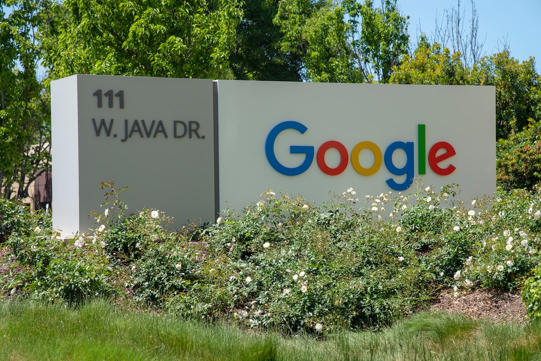 Sign at Google headquarters reads "111 W. Java Dr." with Google's logo in colorful letters. Surrounded by greenery and white flowers.