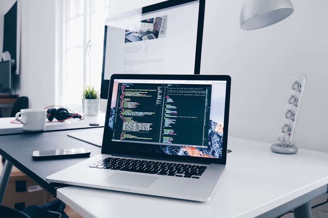 A laptop displaying code is on a desk with a monitor, headphones, a coffee cup, and a lamp.