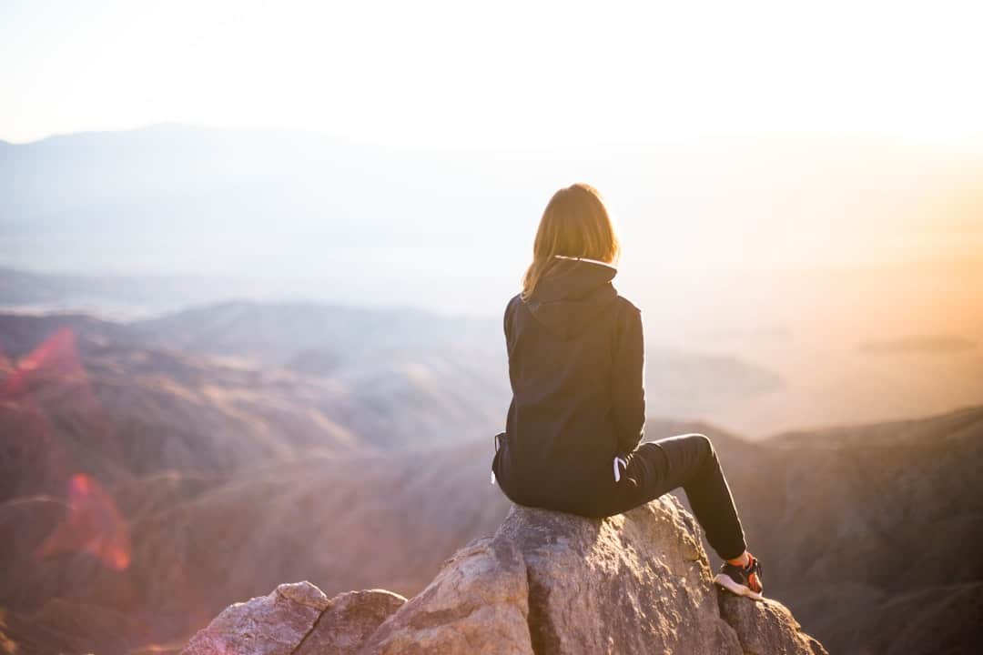 Person in a black jacket sits on a rock, overlooking a sunlit mountainous landscape.