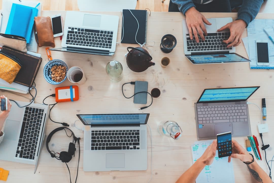 Top-down view of a workspace with five laptops, smartphones, headphones, and assorted stationery items on a wooden table. People are working and using electronic devices.