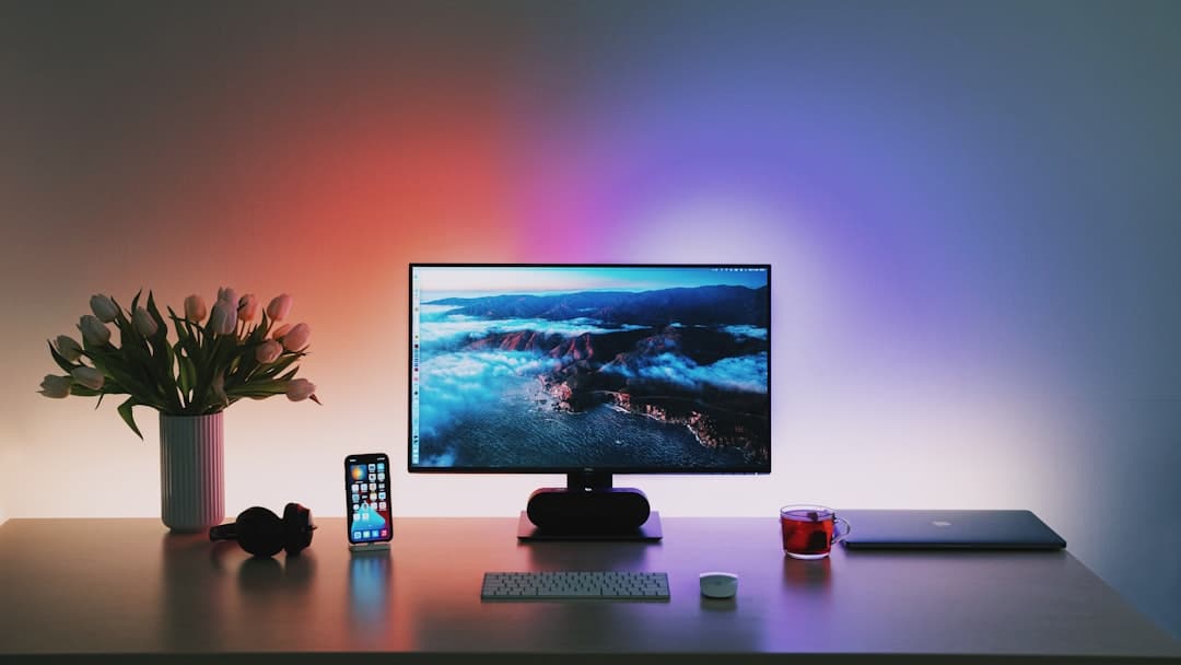 A desk with a computer monitor displaying a scenic image. Items include a smartphone, keyboard, headphones, a mug, a laptop, a small plant, and a vase of flowers. Ambient lighting is visible.