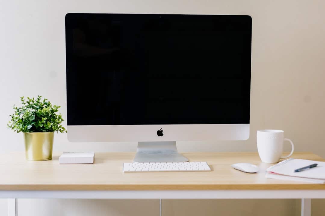 A computer monitor, keyboard, and mouse on a wooden desk, with a potted plant, a cup, and a notebook nearby.