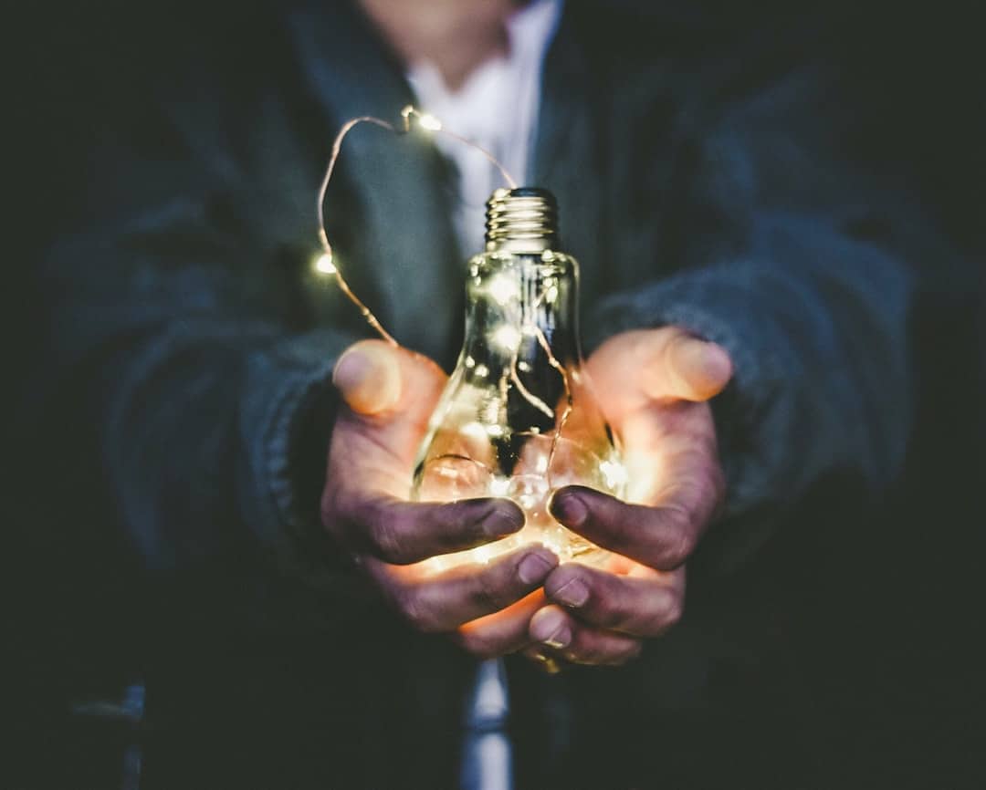 Hands holding a glowing light bulb with small lights inside, against a dark background.