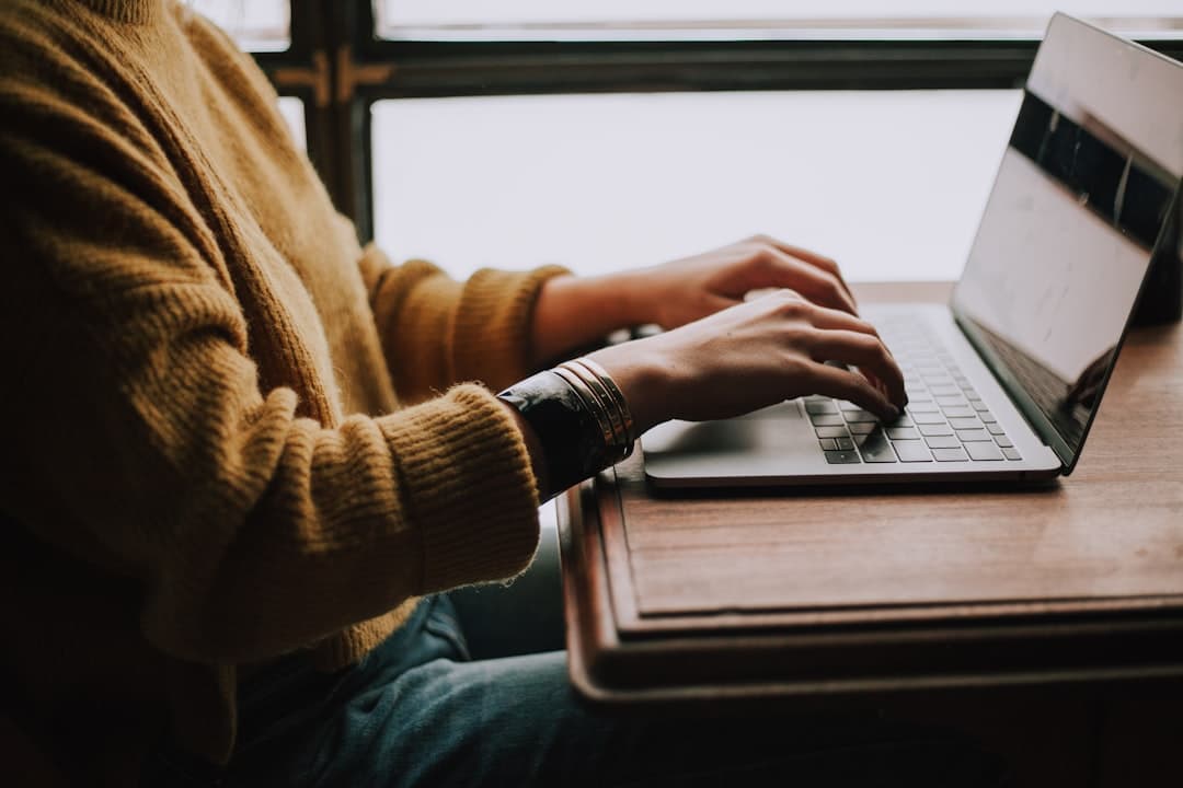 Person in a yellow sweater typing on a laptop at a wooden table, with soft indoor lighting.