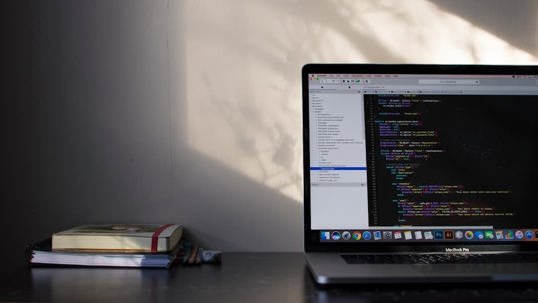 A laptop on a desk displays lines of code on its screen. Beside it are stacked books. Natural light casts shadows on the wall.