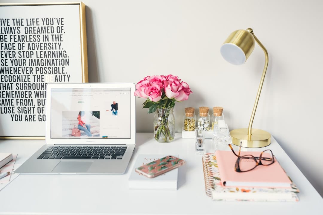 A neatly arranged desk with a laptop, pink flowers in a vase, a gold desk lamp, notebooks, glasses, spices in jars, and a framed inspirational poster.