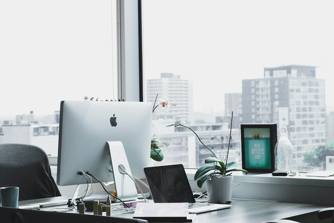 A modern office workspace with an Apple desktop, a laptop, and an orchid plant on a desk near a large window overlooking city buildings.