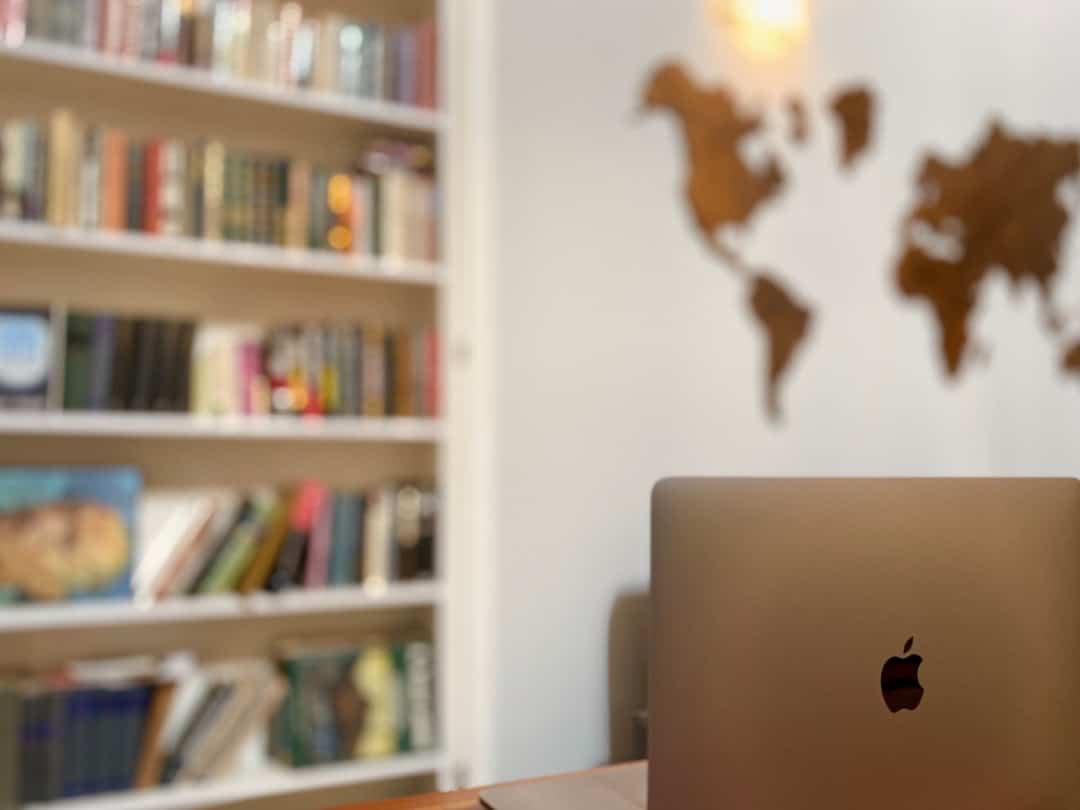 A laptop on a desk in a room with a world map on the wall and a bookshelf filled with books in the background.