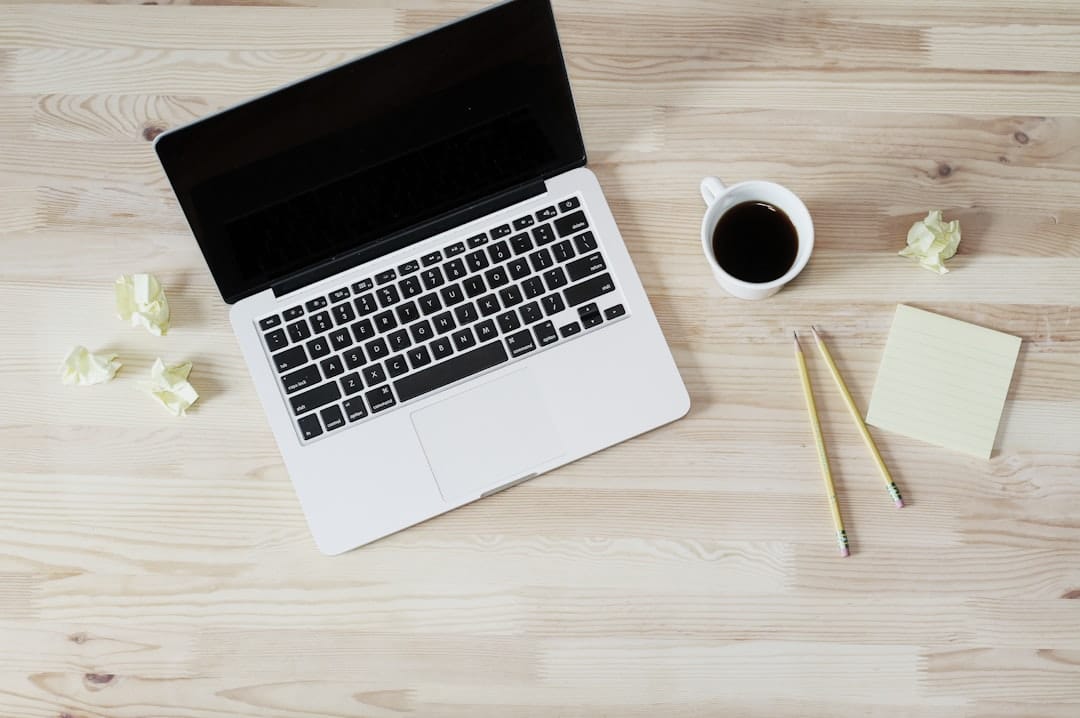 A laptop on a wooden desk with a cup of coffee, two pencils, crumpled paper balls, and a sticky note pad.