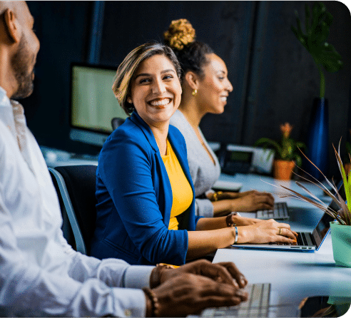 Three people are sitting at a desk with laptops, smiling as they work on hosting solutions in a lively office setting.