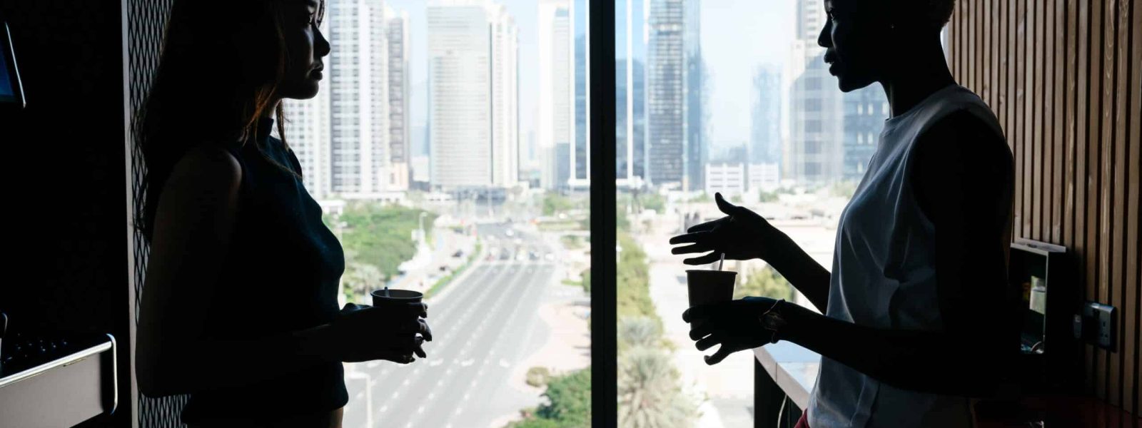 Two people are silhouetted against a large window overlooking a cityscape, holding cups and engaged in conversation.