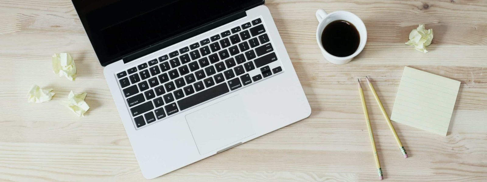 A laptop on a wooden desk with a cup of coffee, two pencils, crumpled paper, and a notepad.