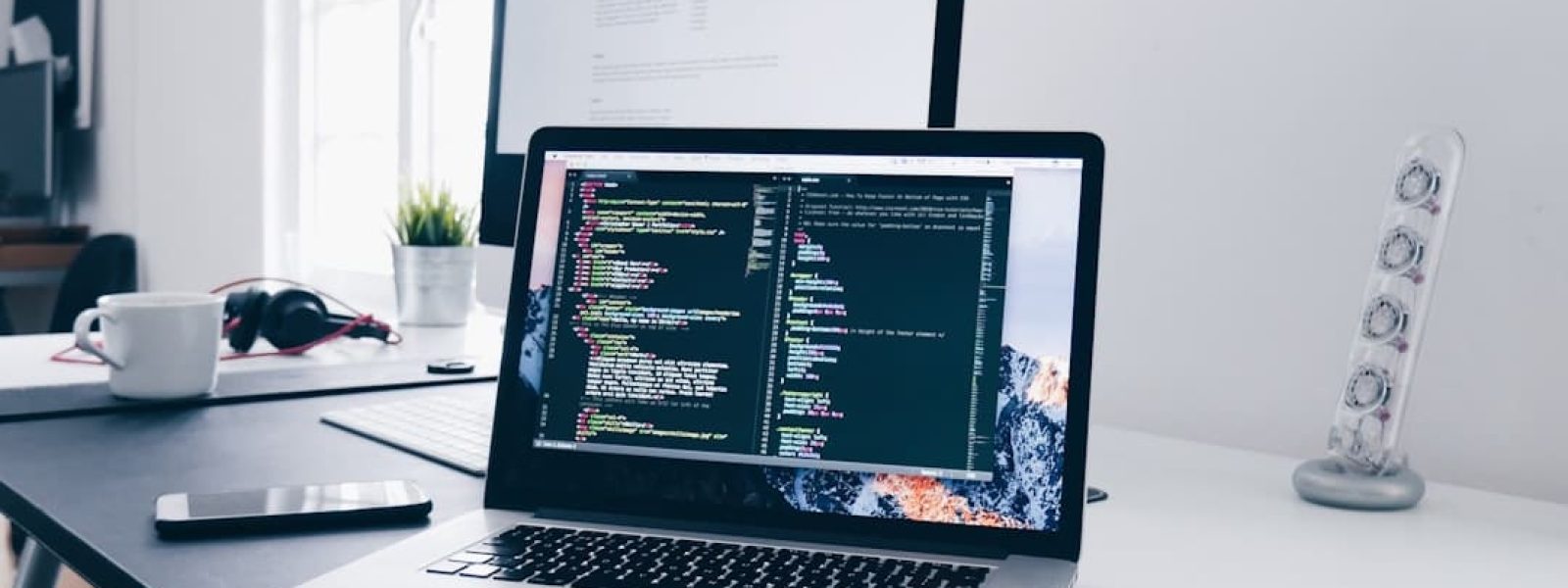 A laptop displaying code is on a desk with a monitor, headphones, a coffee cup, and a lamp.