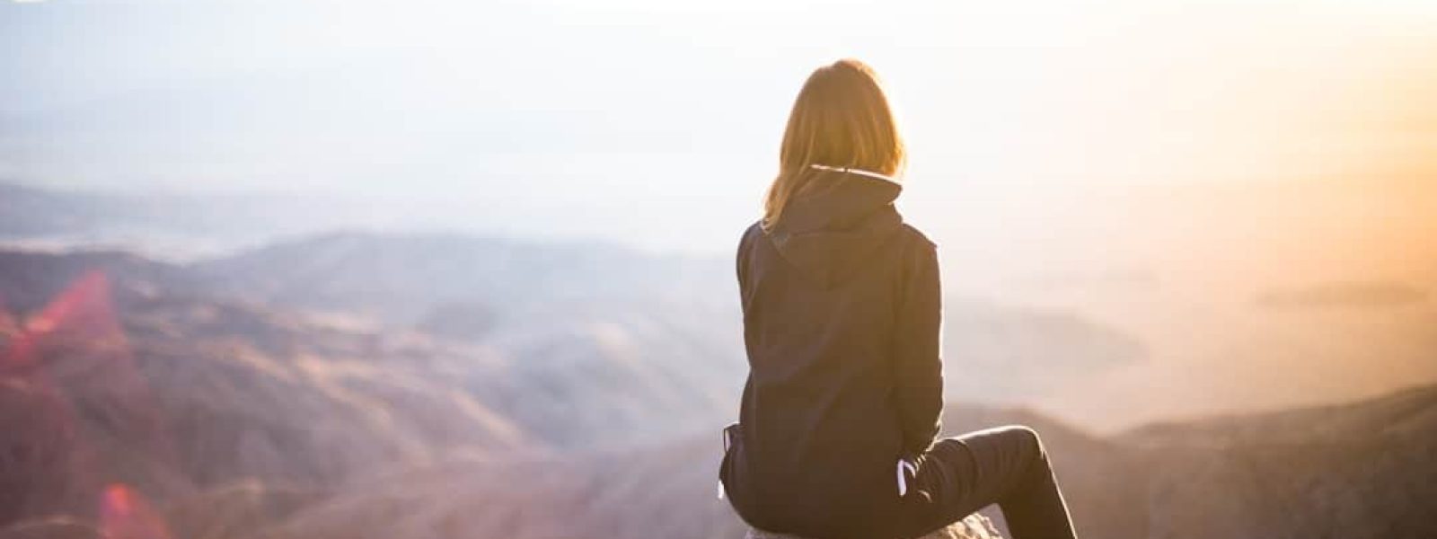 Person in a black jacket sits on a rock, overlooking a sunlit mountainous landscape.