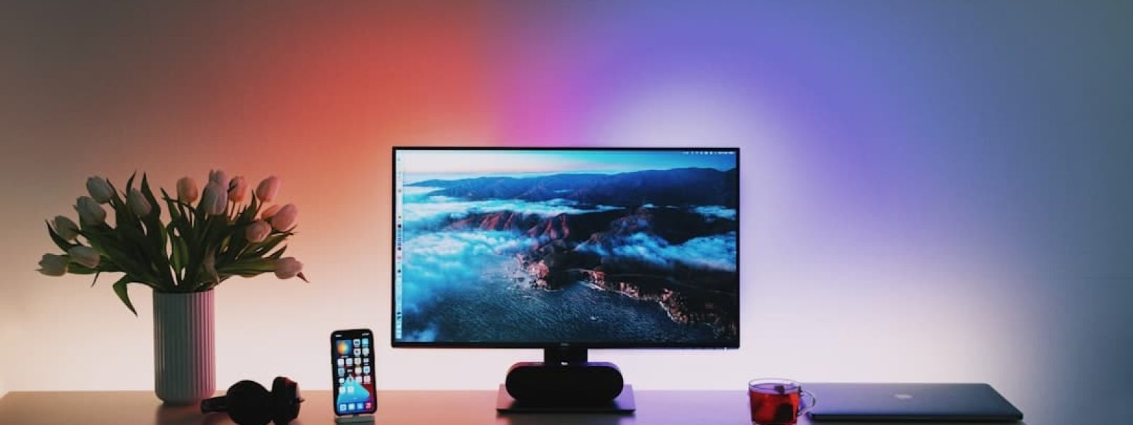 A desk with a computer monitor displaying a scenic image. Items include a smartphone, keyboard, headphones, a mug, a laptop, a small plant, and a vase of flowers. Ambient lighting is visible.