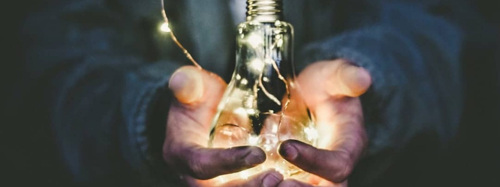 Hands holding a glowing light bulb with small lights inside, against a dark background.