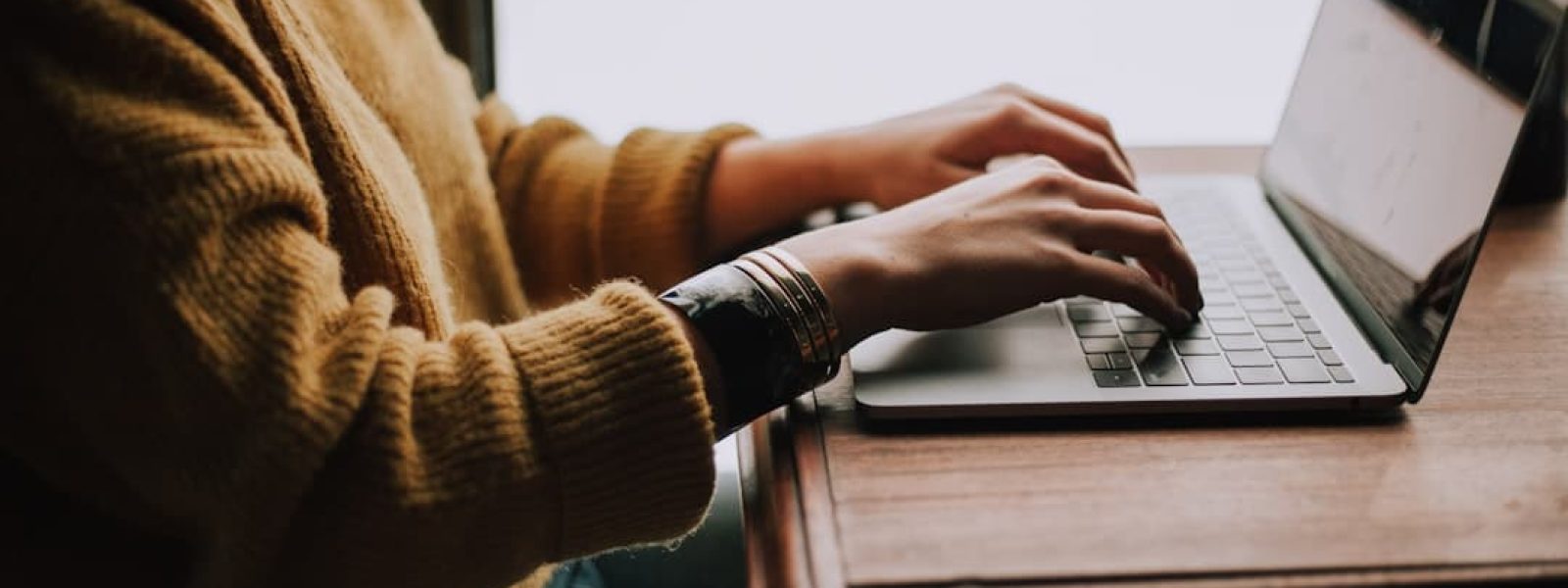 Person in a yellow sweater typing on a laptop at a wooden table, with soft indoor lighting.