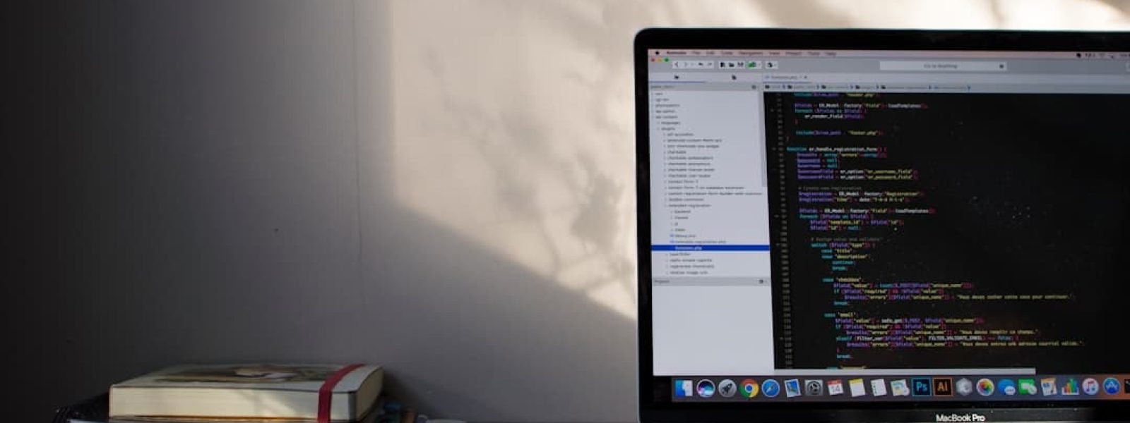 A laptop on a desk displays lines of code on its screen. Beside it are stacked books. Natural light casts shadows on the wall.