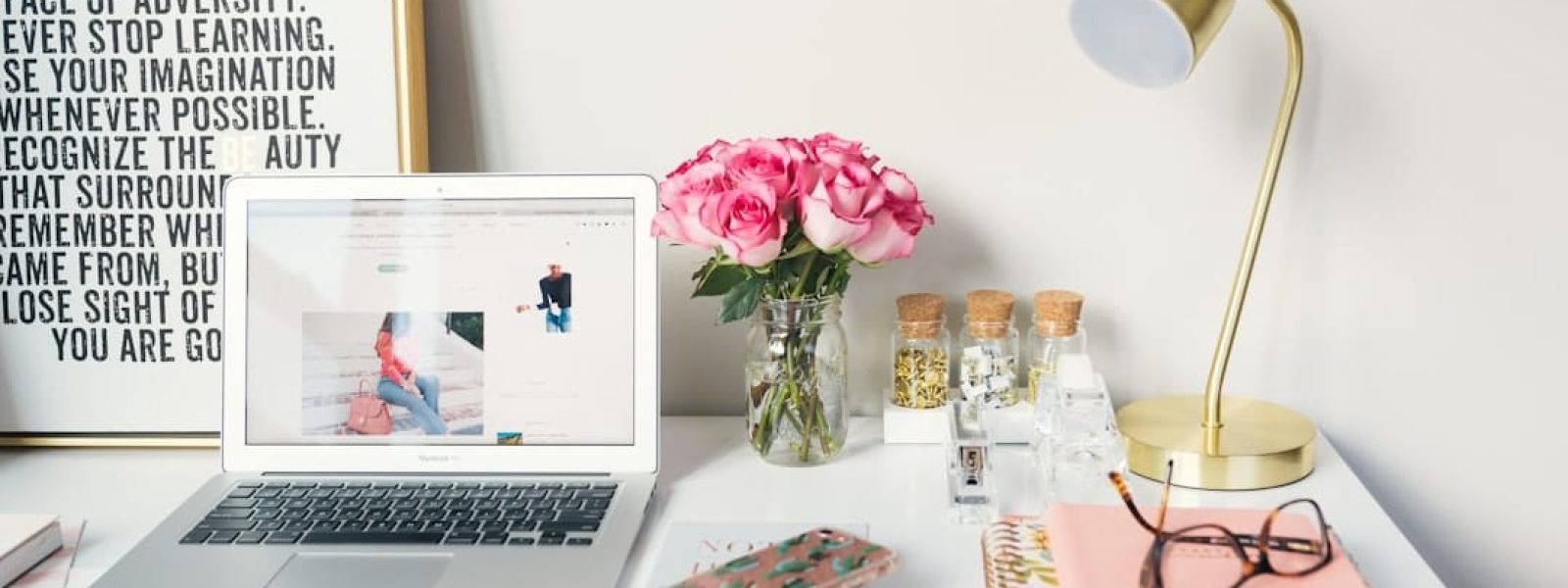 A neatly arranged desk with a laptop, pink flowers in a vase, a gold desk lamp, notebooks, glasses, spices in jars, and a framed inspirational poster.