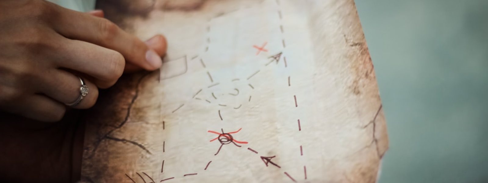 Hands holding an old, weathered treasure map with red X marks, arrows, and dotted paths.