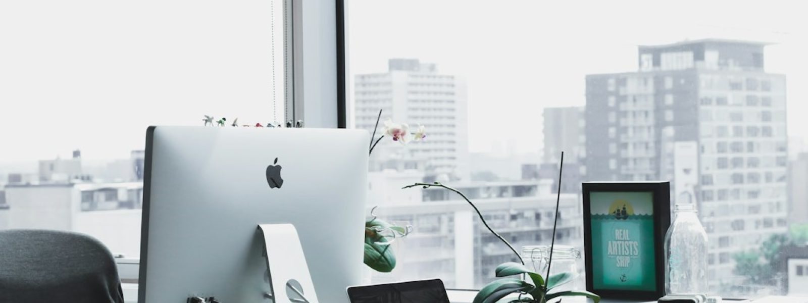 A modern office workspace with an Apple desktop, a laptop, and an orchid plant on a desk near a large window overlooking city buildings.