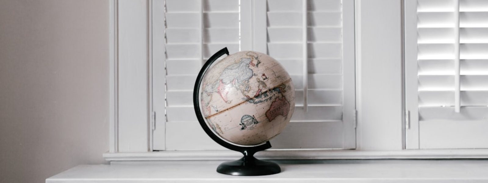 A vintage globe sits on a white radiator cover in front of white window shutters.