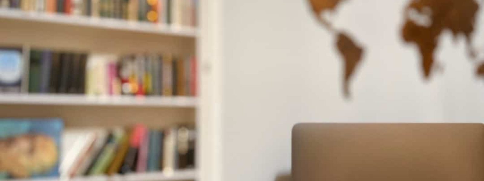 A laptop on a desk in a room with a world map on the wall and a bookshelf filled with books in the background.