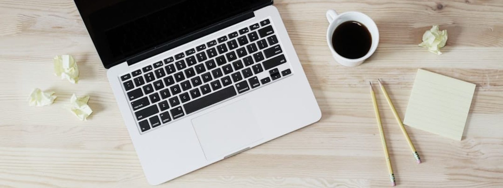 A laptop on a wooden desk with a cup of coffee, two pencils, crumpled paper balls, and a sticky note pad.