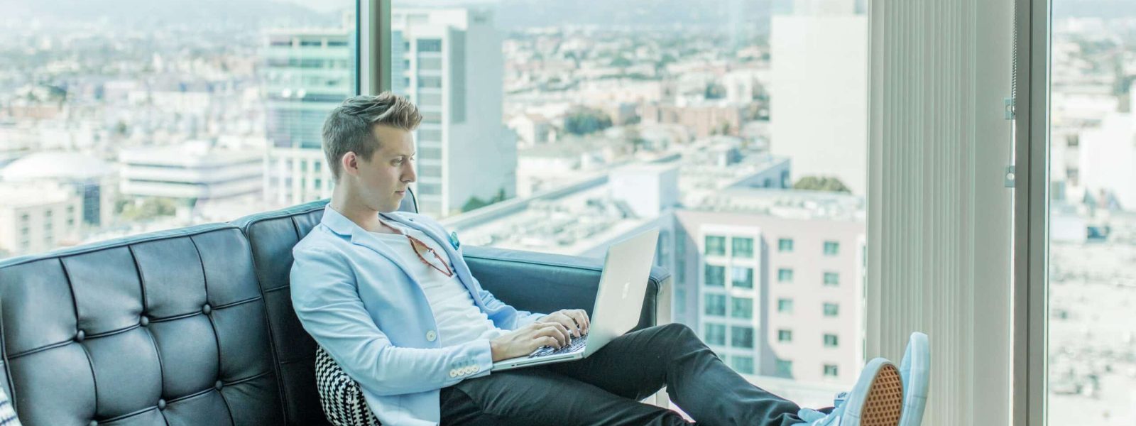 A person sits on a black leather couch, using a laptop, with a cityscape visible through large windows in the background.