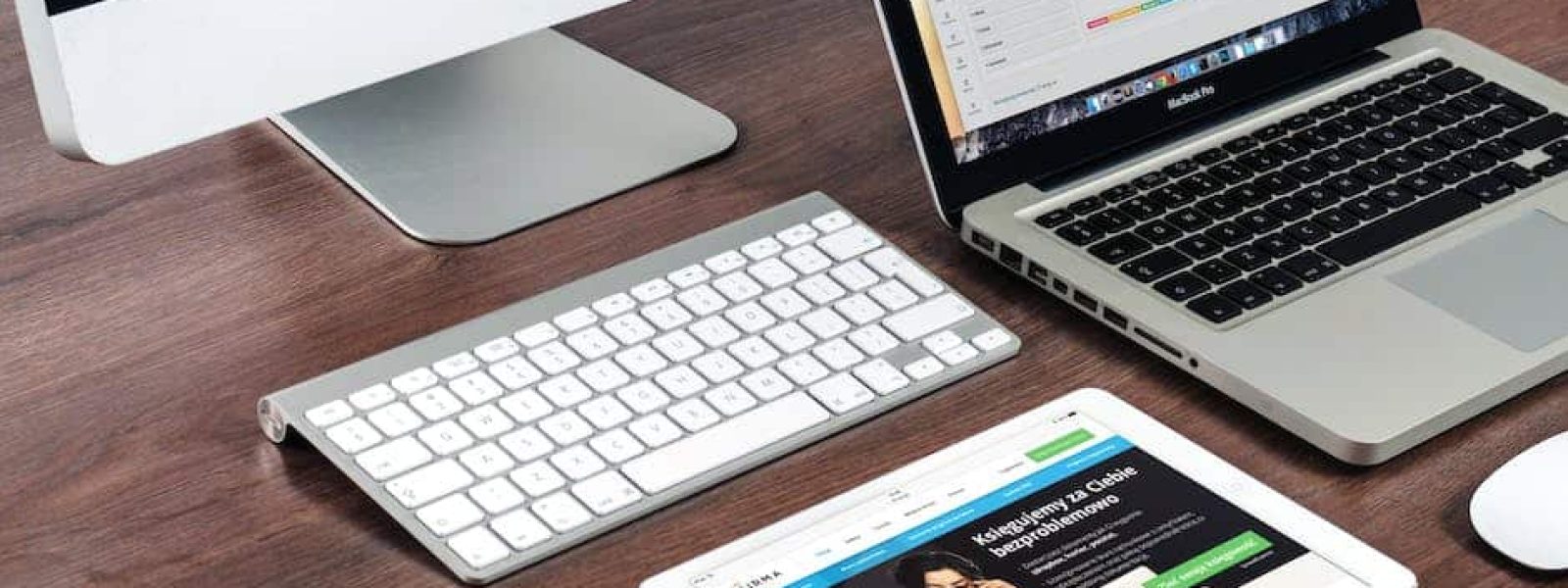 A desk with an iMac, a MacBook, a wireless keyboard, and an iPad displaying a website.