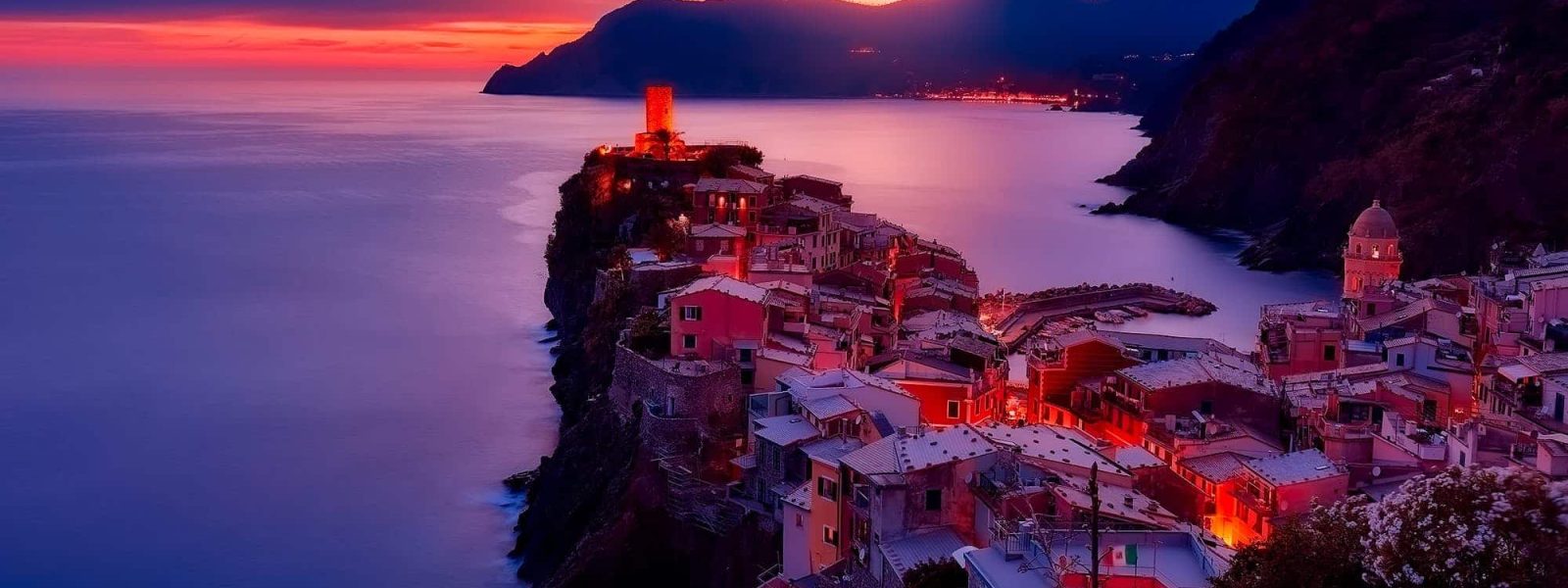 Coastal village on a cliff at sunset, with colorful houses and illuminated streets. The sea is calm, and the sky is a gradient of orange and purple hues.