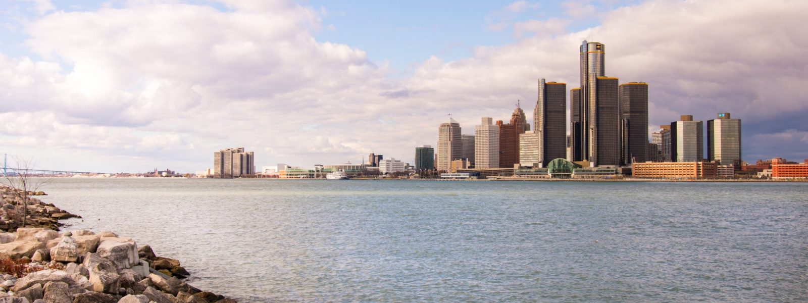 Skyline of a city with tall buildings across a body of water, rocky shoreline in the foreground, under a partly cloudy sky.