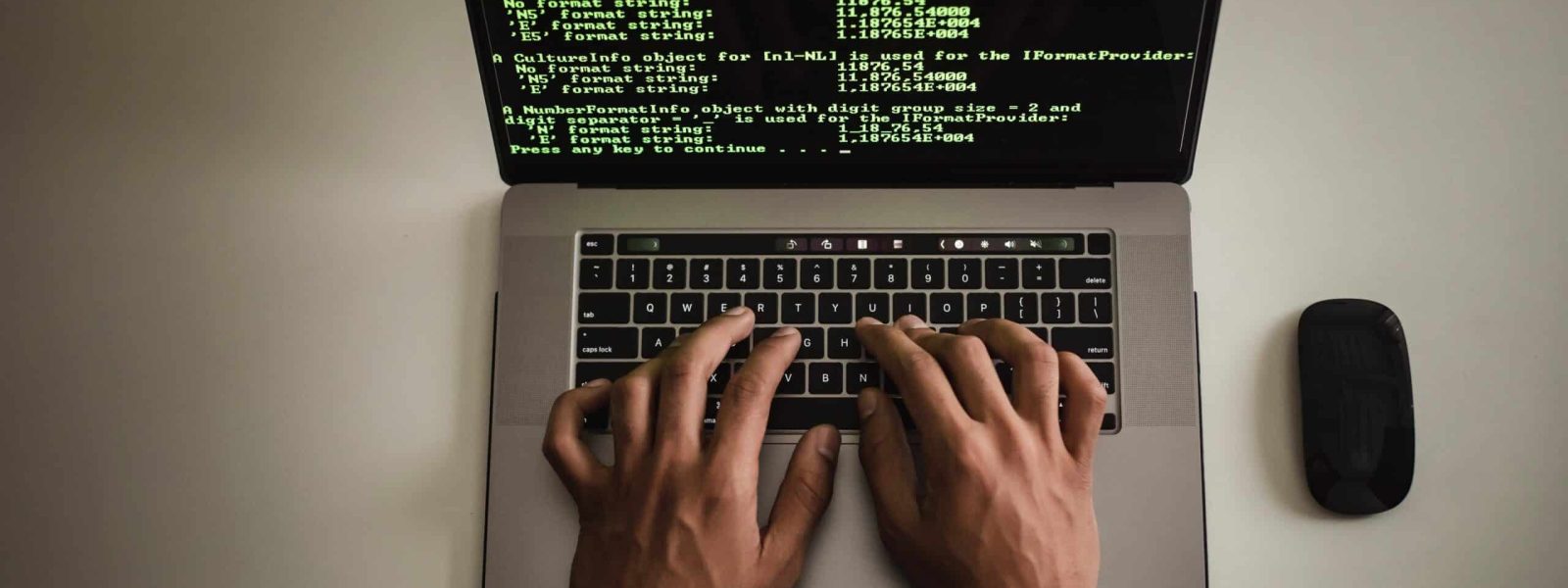 Person typing on a laptop displaying code in a dark-themed text editor, with a black mouse beside it on a white surface.