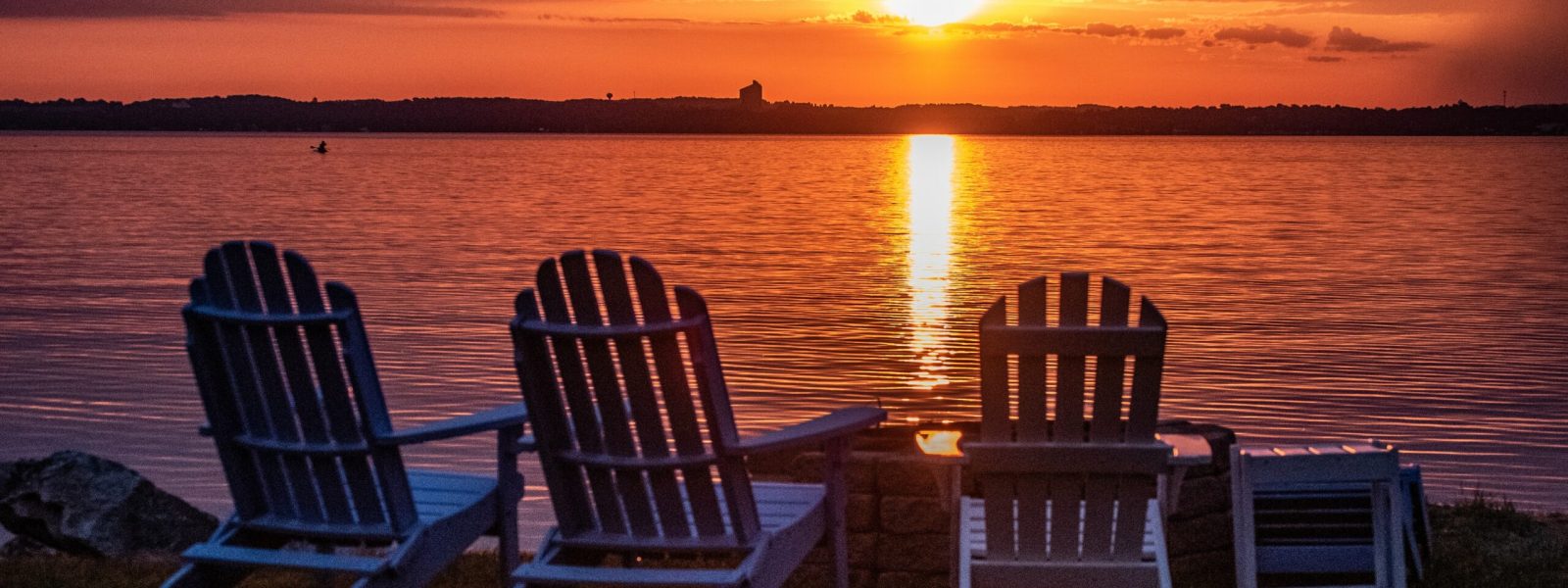 Four Adirondack chairs face a lake under a glowing sunset, with the sun's reflection shimmering on the water.