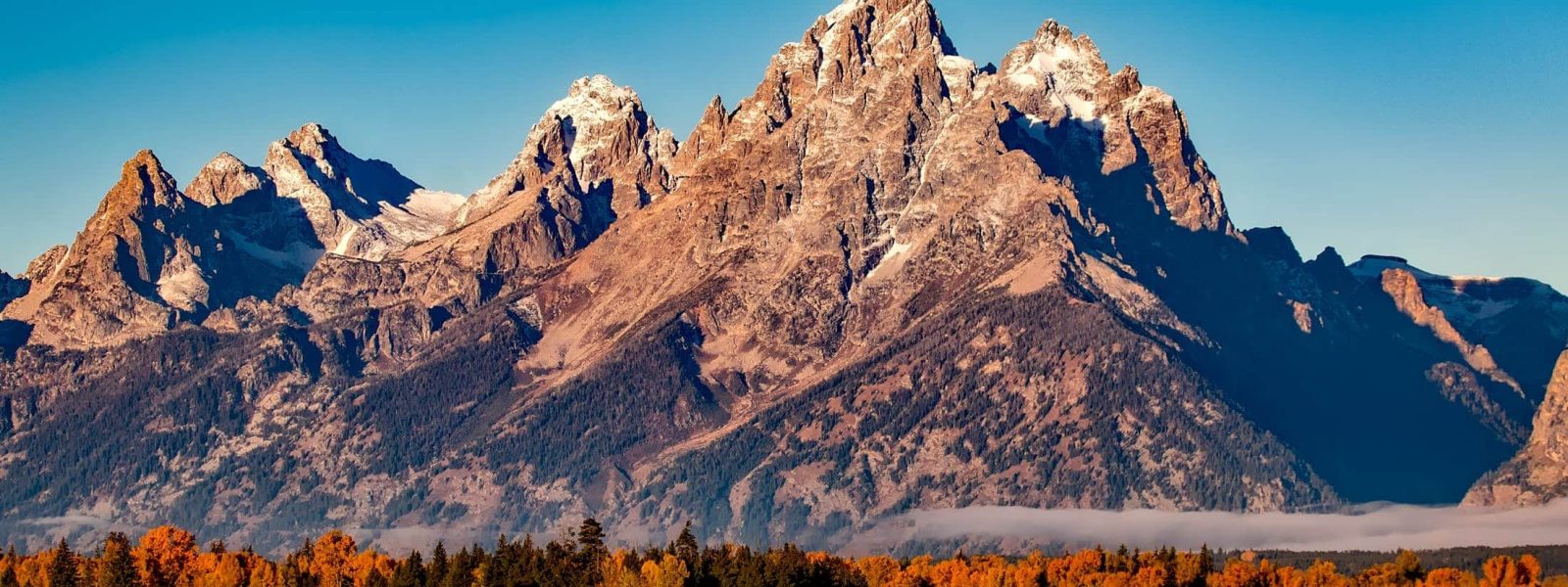 Mountain range with rocky peaks under a clear blue sky, surrounded by an autumnal landscape with trees and open fields in the foreground.